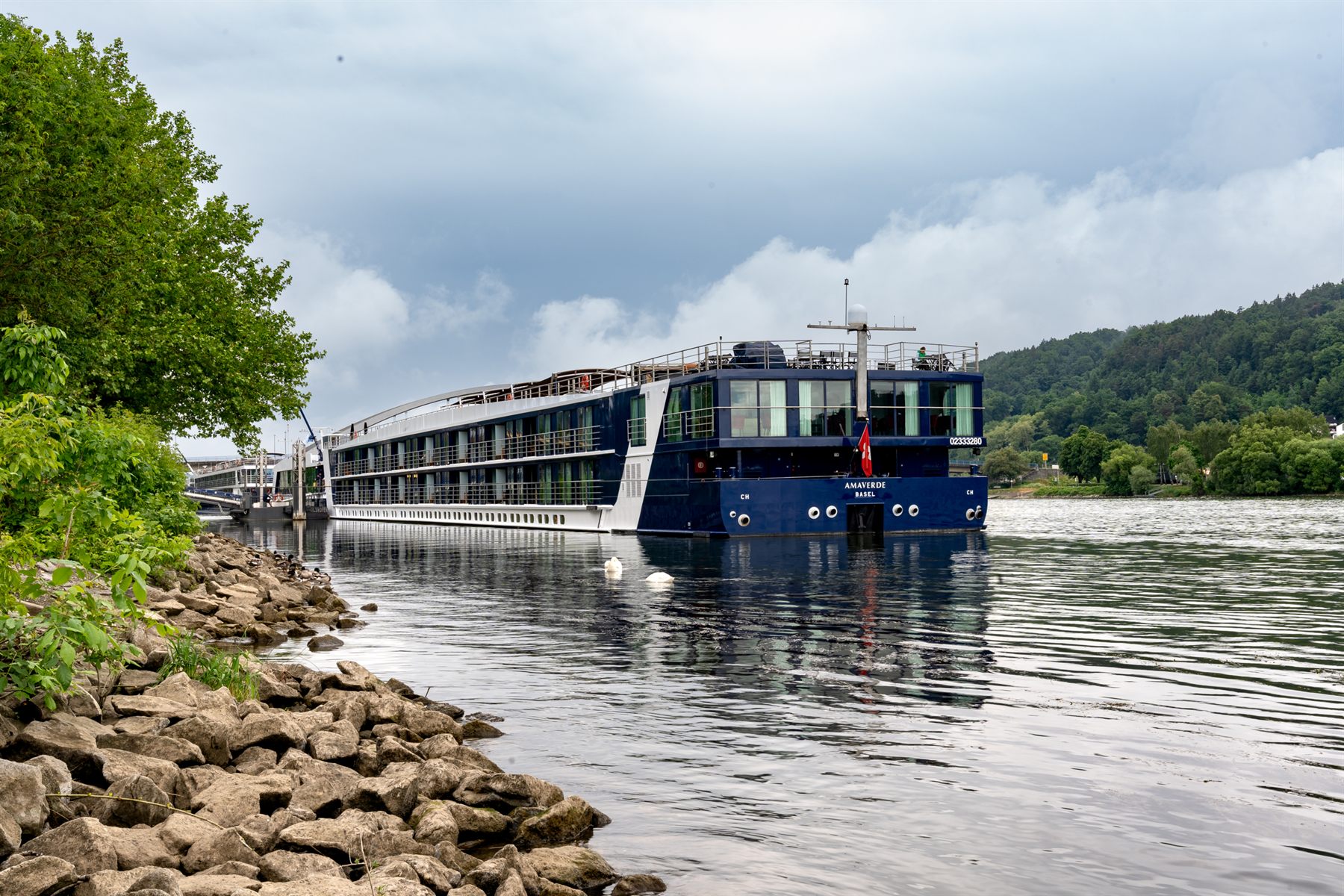 Dark-hulled river cruise ship moored beside a rocky, wooded bank